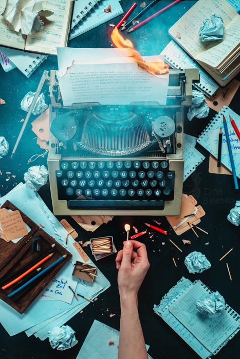 Overhead shot of a typewriter and messy paraphernalia on dark background, an outstretched hand holds a lit match - still life photography ideas.still life photography ideas